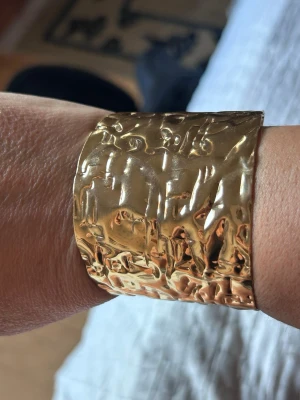 Brett guldigt armband med hamrad yta - Säljer ett brett armband i metall med guldig färg och cool hamrad struktur. Armbandet har en öppen design som gör det enkelt att ta på och av. Perfekt för dig som gillar statement-smycken och vill sticka ut med något unikt.