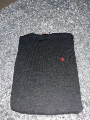 Mörkgrå stickad tröja med röd detalj - Mörkgrå stickad tröja med rund hals och ribbade muddar. På bröstet finns en liten röd broderad logga som ger en snygg kontrast. Tröjan har en klassisk passform och är perfekt för lager på lager. Materialet känns mjukt och bekvämt.