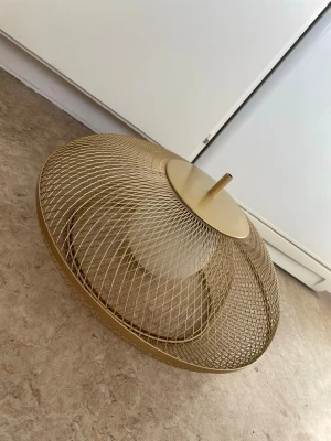 Guldfärgad nätlampa i metall - Säljer en snygg taklampa med guldfärgad metallnät-design. Lampan har rund form och ett stilrent utseende med ett fäste upptill för montering. Perfekt som statement piece i hemmet för dig som gillar modern inredning.