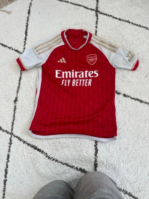 Arsenal matchtröja från Adidas - Säljer en röd och vit Arsenal matchtröja från Adidas med guldfärgade detaljer och logga. Tröjan har klubbemblem på bröstet, sponsortryck och texten 'Visit Rwanda' på ärmen. Materialet är lätt och andas, perfekt för fotboll eller träning.Storlek 11-12 152
