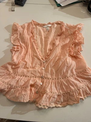 Persikofärgad blus med volanger H&M - Söt persikofärgad blus från H&M med korta ärmar och volangdetaljer. Blusen har knappar framtill, v-ringning och dekorativa spetsband längs volangerna. Materialet är lätt och luftigt, perfekt för en trendig look.