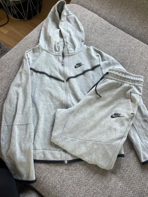 Grå Nike loungewear set med hoodie - Set från Nike i ljusgrå färg. Setet består av en hoodie med dragkedja och matchande mjukisbyxor med elastisk midja och snörning. Väl använd, hoodien är i bättre skick än byxorna, se bilder. Strl XL 158/170