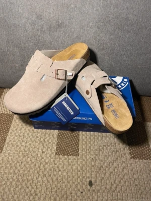 Grå  Birkenstock tofflor i mocka - Säljer ett par snygga grå  tofflor från Birkenstock i mjuk mocka. Modellen har en justerbar rem med metallspänne och klassisk korksula för extra komfort. Perfekta för dig som gillar stilrena och bekväma skor.