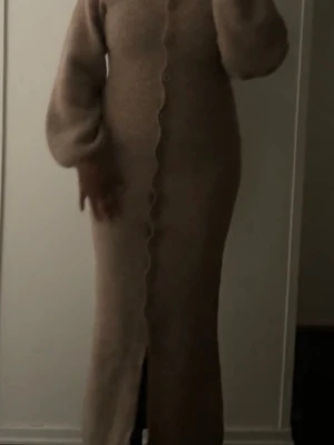 Ginatricot stickad klänning  - Mysig beige stickad långklänning med v-ringning och stora ballongärmar. Klänningen har knappar längs hela framsidan och en slits nedtill. Perfekt för dig som gillar en avslappnad men ändå stilren look.