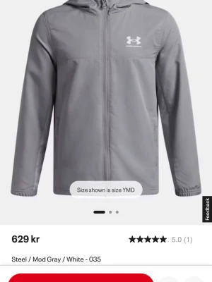 Grå vindjacka från Under Armour - Snygg grå vindjacka från Under Armour med vit logga på bröstet. Jackan har huva, dragkedja framtill och två sidofickor. Materialet är lätt och tåligt, perfekt för att skydda mot vind. Enkel och stilren design som passar till många outfits.