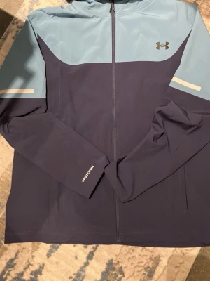 Blå vindjacka från Under Armour - Snygg tvåfärgad vindjacka från Under Armour i mörkblått och ljusblått. Jackan har huva, hel dragkedja framtill och diskreta reflexdetaljer på ärmarna. Tillverkad i lätt polyester, perfekt för aktiva dagar utomhus.