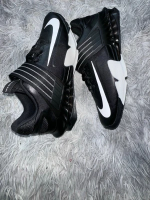 Nike svartvita sneakers med rem - Svarta och vita sneakers från Nike med stor vit swoosh på sidan och sulan. Skorna har en bred rem med vita detaljer över vristen och en robust, mönstrad sula. Materialet är en mix av syntet och textil, med sportig och modern design.