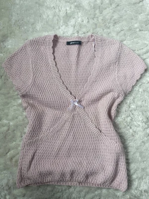 Ljusrosa stickad topp från Gina Tricot - Supersöt ljusrosa stickad topp från Gina Tricot med v-ringning och korta ärmar. Toppen har en liten rosett framtill och ett ribbat mönster längs kanterna. Perfekt för dig som gillar en feminin och trendig look.