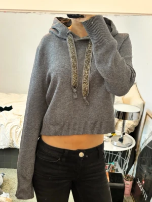 Grå hoodie med glitterband. Zara Knit  - Säljer en grå cropped hoodie med breda glitterband i huvan. Tröjan har ribbade muddar vid ärmslut och nederkant, samt en luva med dekorativa snören. Materialet ser ut att vara mjukt och stickat, perfekt för en avslappnad look men fortafarnde stlish.