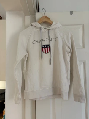 Vit hoodie från GANT med logga - Vit hoodie från GANT med broderad logga och emblem på bröstet. Tröjan har huva med dragsko och långa ärmar. 