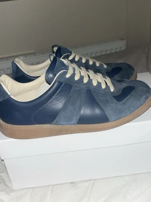 Blå sneakers i mocka och läder - Snygga sneakers i blå mocka och blått läder med beigea skosnören och gummisula. Klassisk låg modell som passar perfekt till jeans eller streetwear. Skorna har en stilren design med kontrasterande material och detaljer.