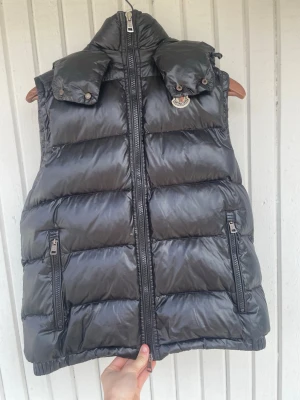Svart dunväst från Moncler - Snygg svart dunväst Moncler med glansig finish, hög krage och avtagbar huva. Västen har två dragkedjeförsedda fickor framtill och Moncler-logga på bröstet. Perfekt för lager-på-lager och streetstyle.