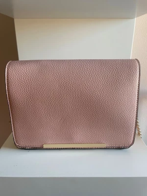 Rosa axelremsväska med guldkedja - Snygg rosa handväska i strukturmönstrat konstläder med guldfärgade detaljer och kedjerem. Väskan har en stilren, rektangulär form och magnetlås. Perfekt för dig som vill ha en trendig accessoar som sticker ut.