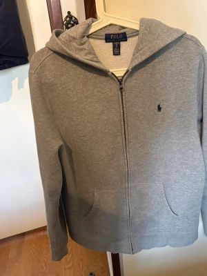 Grå zip hoodie från Polo Ralph Lauren - Snygg grå hoodie från Polo Ralph Lauren med dragkedja framtill och klassisk huva. Tröjan har två fickor och den ikoniska lilla loggan broderad på bröstet. Tillverkad i mjukt bomullsmaterial som är skönt att ha på sig. Perfekt för en avslappnad och stilren look. Tröjan är i storlek 18/20 men motsvarar s väldigt fin tröja. Den är väldigt bra skick.