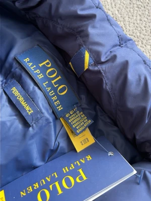 Blå dunväst från Polo Ralph Lauren - Snygg blå dunväst från Polo Ralph Lauren med quiltad design och dragkedja framtill. Västen har ståkrage och den klassiska röda loggan broderad på bröstet. Perfekt att slänga över en hoodie eller tröja för en sportig look.