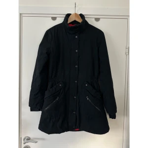 Sisters Point - svart jacka - storlek M - Svart jacka från Sisters Point i storlek M. Jackan har en röd insida med tryck, dragkedjor på fickorna och knappar framtill. Den har även en dragsko i midjan för justerbar passform, samt ”dubbla ärmar” vid ärmsluten för att hålla värmen. Mycket gott skick, som ny. 