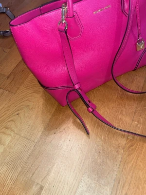Rosa handväska från Michael Kors (ny skick) - Snygg rosa handväska från Michael Kors i kraftigt läder med guldfärgade detaljer och justerbar axelrem. Väskan har en strukturerad form och diskret logga framtill. Perfekt för dig som vill ha en färgklick till din outfit. 100% äkta samt ny skick.
