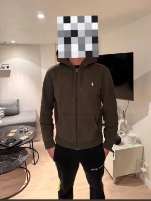 Olivgrön hoodie från Polo Ralph Lauren - Snygg olivgrön hoodie från Polo Ralph Lauren med dragkedja och huva. Klassisk logga broderad på bröstet, ribbade muddar och fickor framtill. Perfekt för dig som gillar stilrena och bekväma plagg. Materialet är mjukt och känns skönt mot huden.