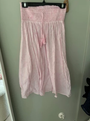 Ljusrosa långkjol med smock och knyt - Söt ljusrosa långkjol med bred smock i midjan och dekorativt knytband med tofsar framtill. Kjolen har en luftig och flowy passform, perfekt för varma dagar. Tillverkad i ett mjukt och tunt material som ger en avslappnad känsla.