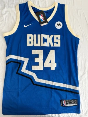 Blå Milwaukee Bucks basketlinne Nike - Snyggt blått basketlinne från Nike med Milwaukee Bucks tryck och nummer 34. Antetokounmpo står på ryggen. Linnet har vita och svarta detaljer, breda ärmhål och rund hals. Perfekt för basketträning eller att bära som supporterplagg.