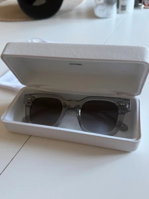 Grå CHIMI 04 solglasögon med box - Snygga solglasögon från CHIMI med transparent grå ram och mörka glas. Modellen har en fyrkantig form och kommer med originalbox, förvaringspåse och putsduk. Perfekt för dig som vill ha en stilren och modern look.