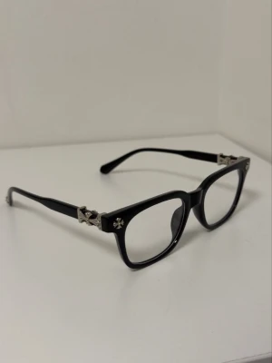 Black Chrome Hearts Bluelight Sunglasses - Säljer ett par svarta solglasögon från Chrome Hearts med rektangulära bågar och coola silverkors på sidorna och ändarna. Glasögonen har en klassisk och edgy look med tydliga metallinslag som sticker ut. Perfekt för dig som vill ha något unikt och stilrent.