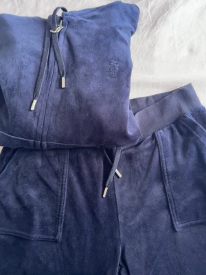 Mörkblå velourset från Juicy Couture - Mysigt mörkblått loungewear-set från Juicy Couture i velour. Setet består av en zip-hoodie med dragsko och matchande byxor med fickor och dragsko i midjan. Diskret Juicy Couture-logga på bröstet och metall-detaljer på snören och dragkedja.