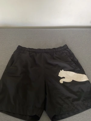 Svarta shorts från Puma med logga -  shorts från Puma med vit logga på både fram- och baksidan. De har elastisk midja och sidofickor, perfekt för sport eller chill. Materialet är syntet och ger en lätt känsla. Klassisk design som funkar till många outfits.