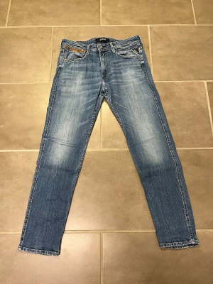 Replay Anbass blå jeans - Säljer ett par blå Replay Anbass jeans med klassisk femficksmodell och raka ben. Jeansen har snygga slitningar och ljus tvätt, samt Replay-logga på fickan. Tillverkade i bomull med en bekväm passform. Perfekta för en avslappnad och trendig stil. 