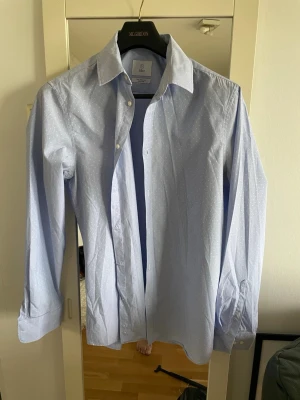 Ljusblå skjorta från bläck  - Klassisk ljusblå skjorta från bläck  med diskret prickigt mönster. Skjortan har lång ärm, knappar framtill och en snygg krage. Tillverkad i bomull för en skön och luftig känsla. Perfekt för dig som gillar stilrena och fräscha plagg.