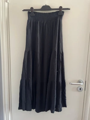 Svart långkjol med smock - Svart långkjol med smockad midja och lätt rynkad, flödande form. Kjolen är enkel och stilren, perfekt för dig som gillar en avslappnad och trendig look. Materialet känns mjukt och luftigt, vilket gör den bekväm att bära under varmare dagar.
