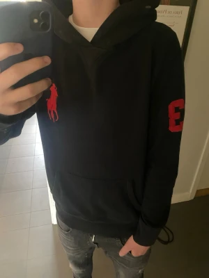 Polo Ralph Lauren Hoodie  - Säljer en riktigt snygg och klassisk hoodie från Polo Ralph Lauren i svart med den ikoniska "Big Pony"-loggan broderad i rött på bröstet. 🔴⚫️ Hoodien har även den klassiska "3"-patchen på höger ärm, vilket ger den en sportig och lyxig look. Detaljer: • Märke: Polo Ralph Lauren 🐎 • Storlek: Medium (M) 📏 • Färg: Svart med röda detaljer. • Modell: Klassisk hoodie med luva och känguruficka framtill. • Material: Mjuk och skön bomullsblandning med hög kvalitet. 