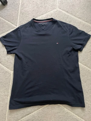 Mörkblå t-shirt från Tommy Hilfiger - Säljer en mörkblå t-shirt från Tommy Hilfiger med klassisk v-ringning och diskret logga på bröstet. T-shirten är gjord i mjuk bomull och har korta ärmar. Perfekt för dig som gillar stilrena plagg med en touch av premium.