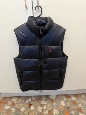 Svart dunväst från Polo Ralph Lauren - Snygg svart dunväst från Polo Ralph Lauren med hög krage och dragkedja framtill. Västen har två sidofickor med dragkedja och en röd logotyp broderad på bröstet. Perfekt för lager-på-lager och håller dig varm under kyliga dagar.