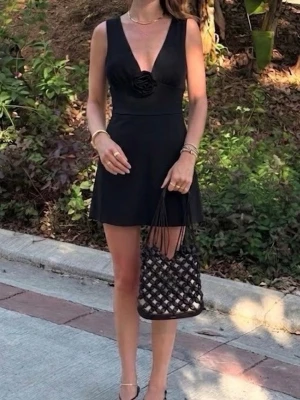 Svart kortklänning med djup v-ringning - Svart kortklänning med djup v-ringning och en dekorativ blomdetalj framtill. Klänningen är ärmlös och har en lätt utsvängd form som ger en snygg siluett. Perfekt för dig som vill ha en stilren och elegant look.