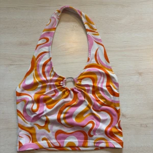 Färgglad halterneck topp med ring - Cool halterneck topp med ringdetalj framtill och retro swirl-mönster i rosa, orange och vitt. Perfekt för dig som gillar färg och unik stil. Toppens form är croppad och materialet känns mjukt och stretchigt.
