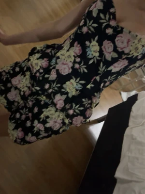 Blommig kort klänning från H&M - Supersöt kort klänning från H&M med svart botten och stora blommor i rosa och gult. Klänningen är ärmlös och har en volangig kjol som ger en lekfull vibe. Perfekt för varma dagar och sommarhäng!