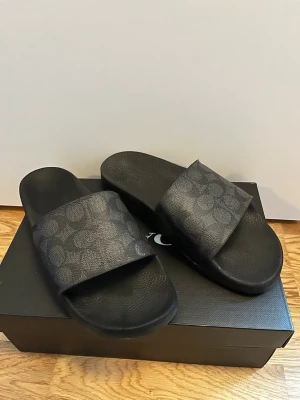 Coach charcoal slides med logomönster - Säljer ett par snygga slides från Coach i färgen charcoal med deras klassiska logomönster på remmen. Sandalerna har en bred, bekväm rem och svart sula. Perfekta för chill dagar och enkel stil.
