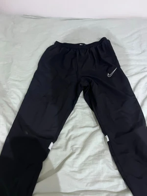 Svarta Nike träningsbyxor - Säljer ett par svarta träningsbyxor från Nike med vit logga på höften och vita detaljer på benen. Byxorna har elastisk midja och är tillverkade i ett lätt och sportigt material som passar perfekt för träning och aktivitet.