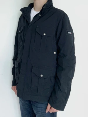Svart utilityjacka från Woolrich - Snygg svart utilityjacka från Woolrich med flera praktiska fickor framtill och tryckknappar. Jackan har hög krage och patch med logga på ärmen. Perfekt för dig som gillar en stilren och funktionell look.