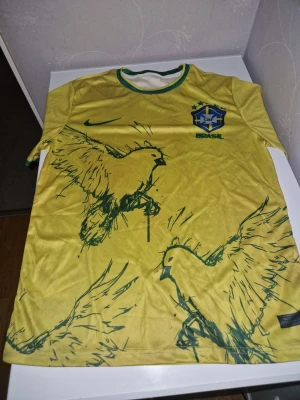Gul Brasil Nike fotbollströja - Snygg gul fotbollströja från Nike med Brasil-logga på bröstet och gröna detaljer. Tröjan har coolt grafiskt tryck med fåglar i grönt och rund hals med gröna och vita ränder. Perfekt för dig som älskar fotboll och vill sticka ut på planen.
