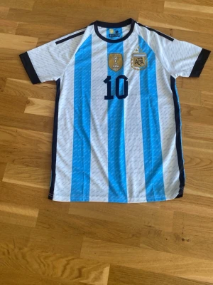 Argentina Messi matchtröja blåvit - Säljer en officiell Argentina fotbollströja från Adidas med Messi och nummer 10 på ryggen. Tröjan har klassiska blå och vita ränder, svarta detaljer vid ärmslut och krage samt broderade märken på bröstet. Perfekt för dig som älskar fotboll och vill stötta Argentina.