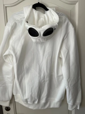 Vit hoodie med goggles från C.P. Company - Liten i storlek så passar dig som har storlek M också. Säljer en vit hoodie från C.P. Company med ikoniska goggles i huvan. Tröjan har dragkedja framtill, ribbade muddar och är tillverkad i mjukt material. 