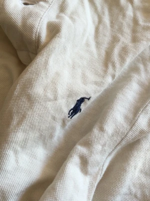 Vit piké  från Polo Ralph Lauren - Vit polotopp från Polo Ralph Lauren med klassisk krage och blå broderad logga på bröstet. Toppens material är bomull och den har en normal passning. Perfekt för dig som gillar stilrena och tidlösa plagg.