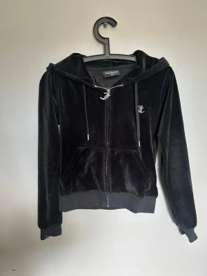 Svart velour hoodie från Juicy Couture - Säljer en svart velour hoodie från Juicy Couture med dragkedja, huva och fickor fram. På ryggen står det 'Juicy' i glittriga stenar och på bröstet finns ett litet JC-emblem. Perfekt för dig som gillar klassisk Y2K-stil.