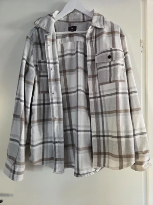 Rutig overshirt från H&M i beige och grå - Snygg overshirt från H&M i mjukt material med rutigt mönster i beige, grått och vitt. Skjortan har två bröstfickor med lock, knappar framtill och klassisk krage. Perfekt lager på lager-plagg för en avslappnad stil.