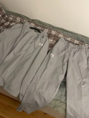 Grått träningsset från Under Armour - Snyggt grått träningsset från Under Armour med jacka och byxor. Setet har meshfoder på insidan för extra ventilation och komfort. Båda plaggen har Under Armour-logga i vitt och är perfekta för träning eller uppvärmning.