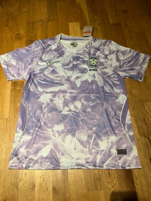 Lila Korea Nike fotbollströja - Säljer en unik fotbollströja från Nike med Koreas landslagsemblem. Tröjan har ett lila och vitt blommigt mönster, korta ärmar och rund hals. Tillverkad i lätt och ventilerande material, perfekt för träning eller match.