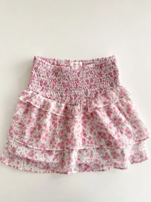 Rosa blommig volangkjol - Supersöt kort kjol med blommigt mönster i rosa och vitt. Kjolen har smock i midjan och dubbla volanger för en extra fluffig look. Perfekt för vår och sommar och passar till både sneakers och sandaler.