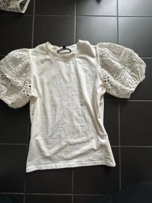 Vit topp med spetsärmar - Trendig vit topp med puffiga, korta spetsärmar och rund halsringning. Kroppen är i mjuk bomull medan ärmarna har ett detaljerat spetsmönster. Toppen sitter nära kroppen💓